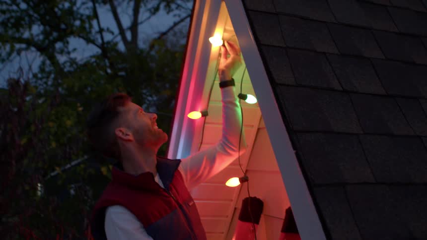 Man smiling as he strings up christmas lights under the eaves at night