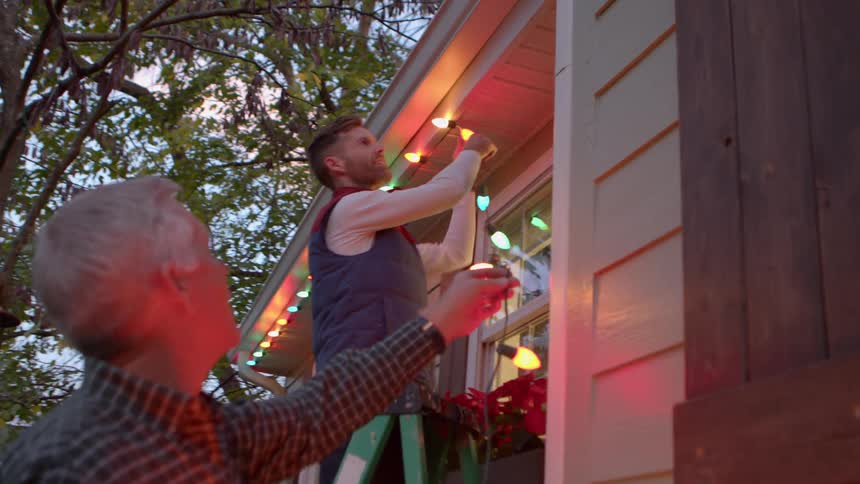 Adult son hanging up christmas lights under the eaves on house as dad holds up the ends