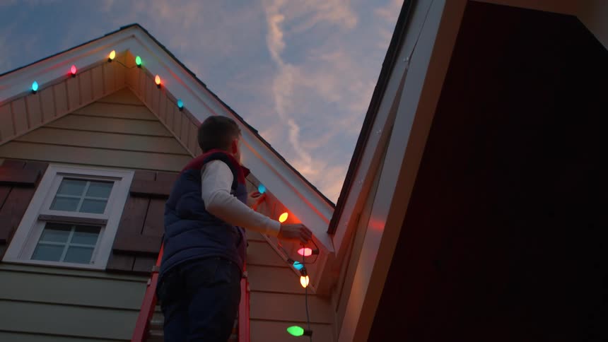 The man is standing on a ladder hanging christmas decorations on his home