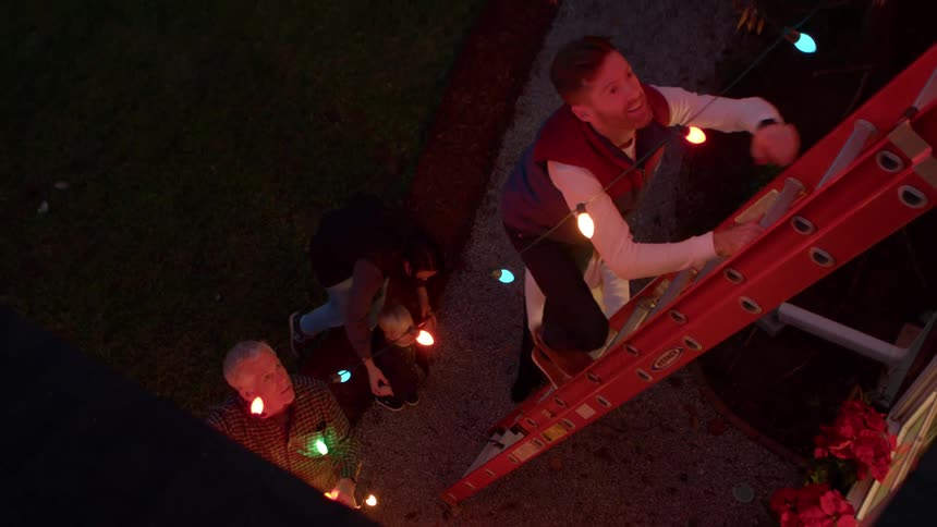 A man climbs a ladder toward the roof to hang christmas lights at night while two women and an elderly man stand on the ground with one of the women playing with a toddler