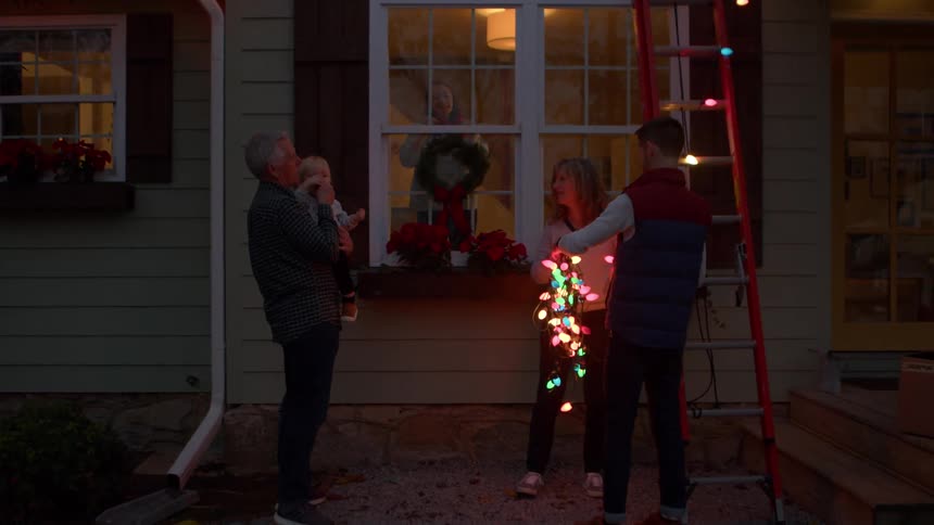 A man climbs a red ladder while holding a string of colorful lights preparing to decorate the roof of a house