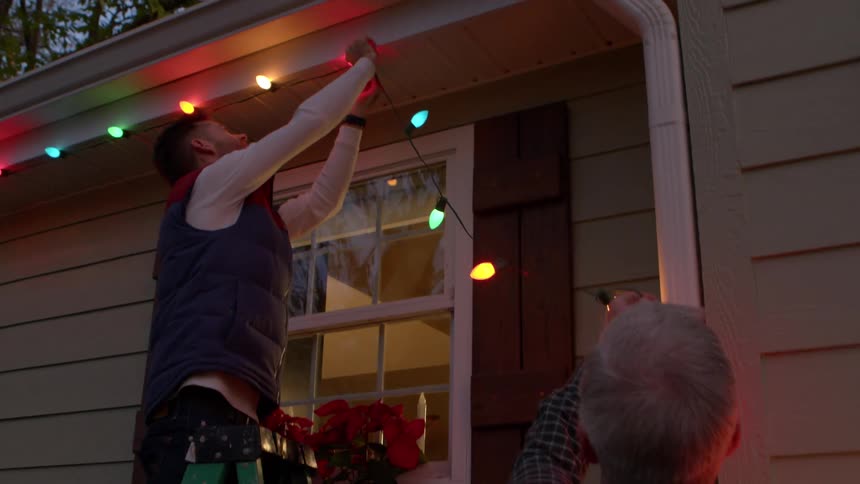 Adult son hanging up christmas lights on house as dad holds up the ends