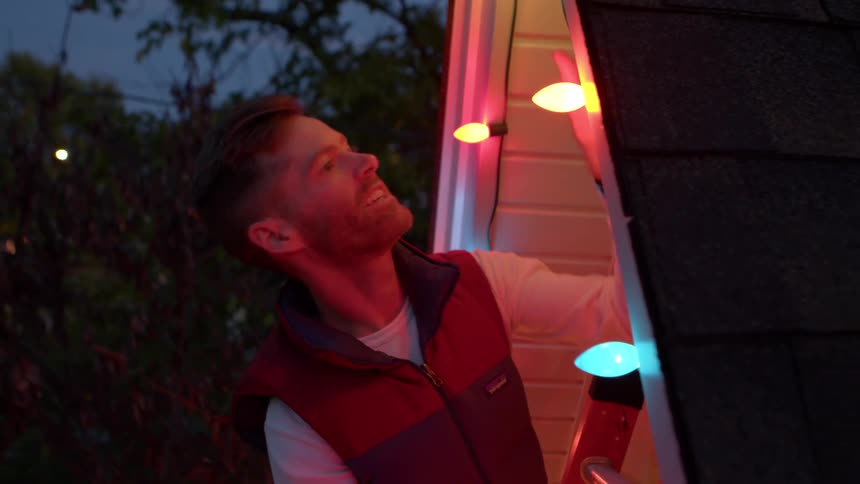 A man in a red and blue vest stands on a ladder hanging colorful lights on the white siding of a building