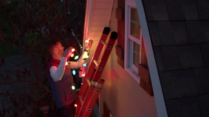 Man decorates house with colorful lights from a ladder during nighttime