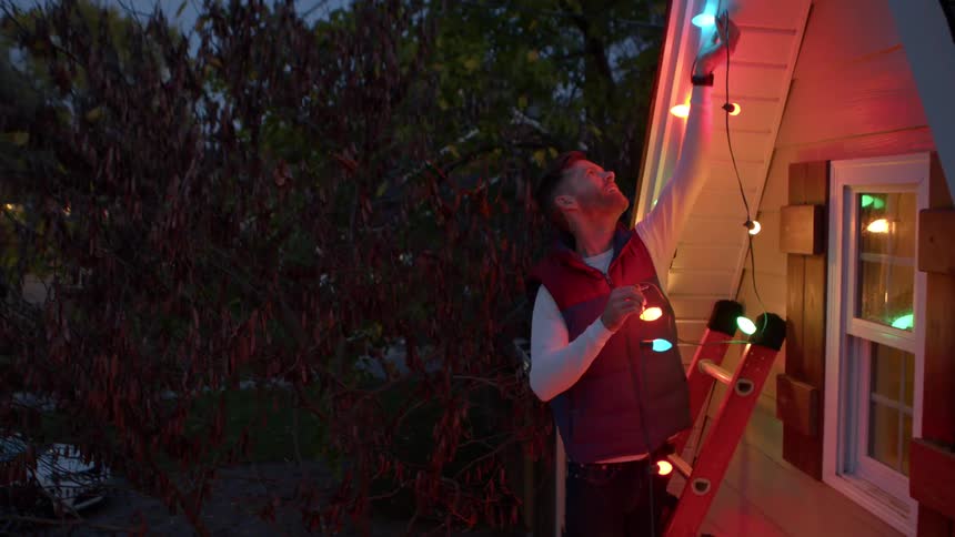 A man stands on a ladder stringing colorful lights along the roofline of a house at dusk
