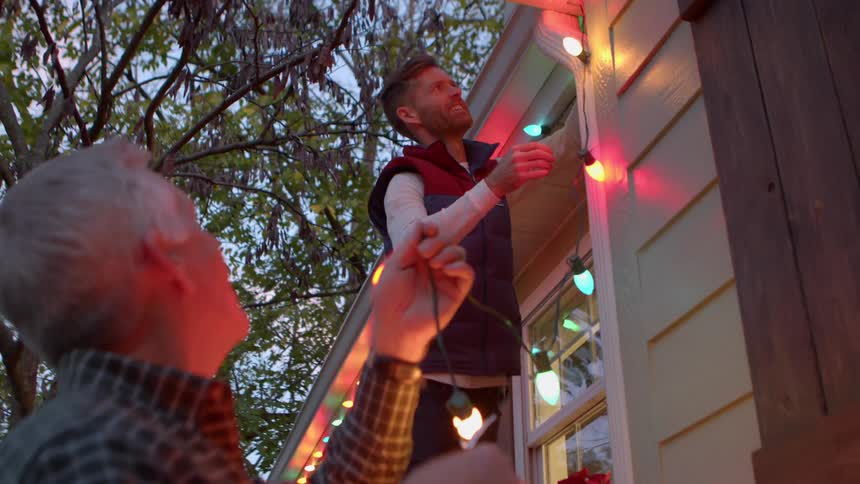 Two men hang colorful string lights outside a house at dusk