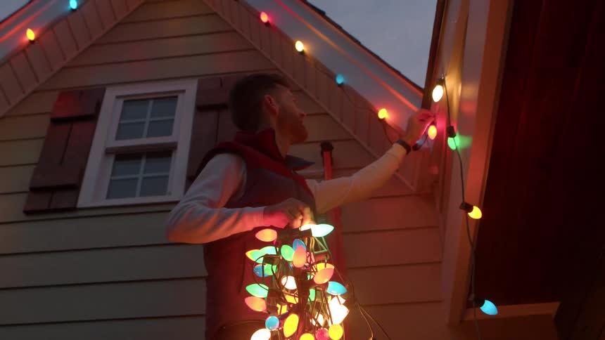 A woman hangs a green wreath inside a window as a man on a red ladder secures colorful string lights to a house eave holding a bundle of lights