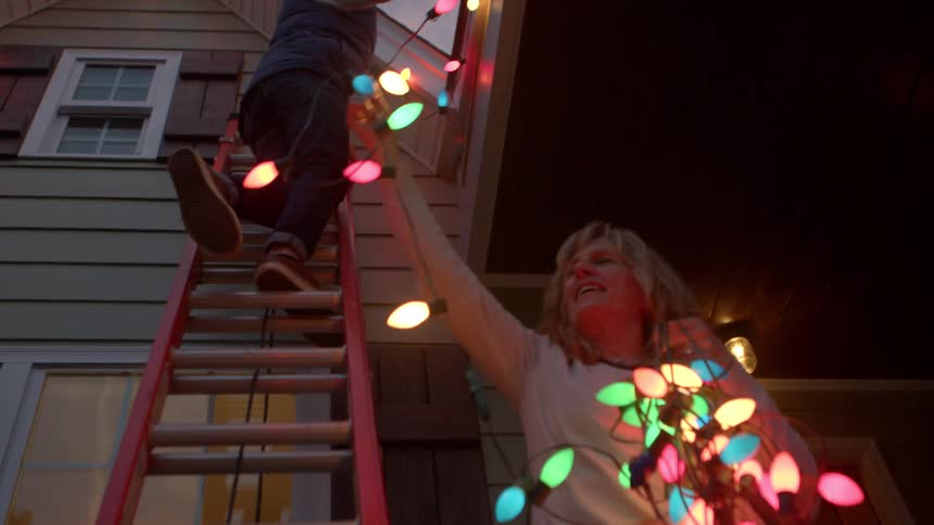 Couple works together to put a string of lit colored lights onto their house after dusk