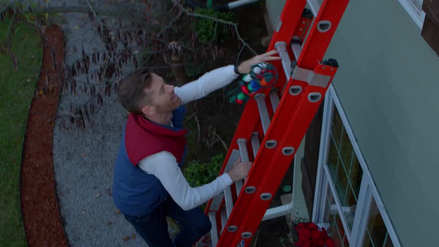 Two men operating a ladder on the outside wall of a house with one of them climbing up with christmas lights