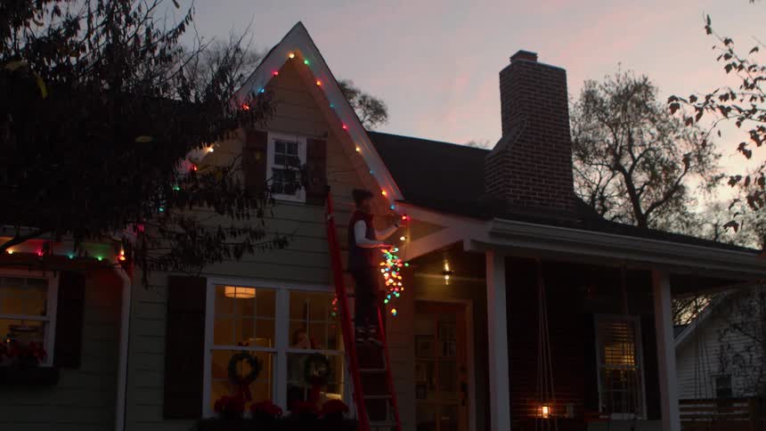 A person on a ladder hangs christmas lights on a house at dusk