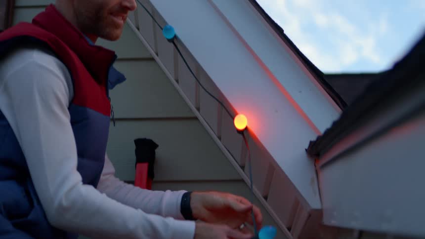 A man hangs colorful string lights on the exterior of a house
