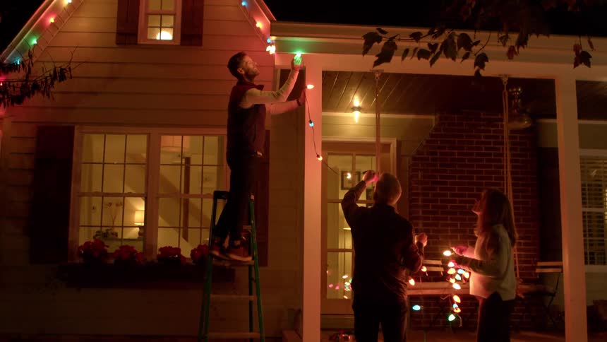 A family decorating the exterior of their house with colorful string lights at night a man is on a ladder attaching lights to the roofline while a man and a woman on the ground hold more strands of lights working together to illuminate their home