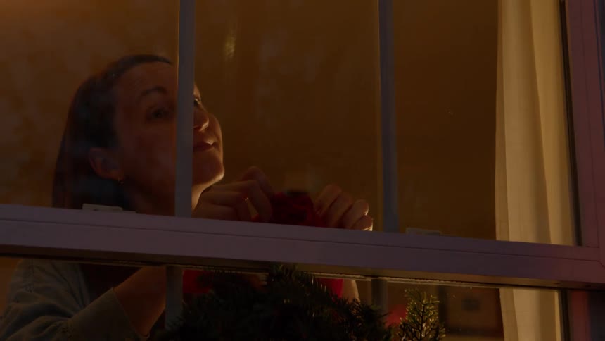 Woman hangs a christmas wreath while smiling at the person hanging christmas lights outside at night