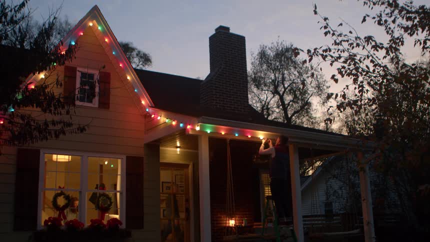 Person hanging colorful lights on house roof at dusk