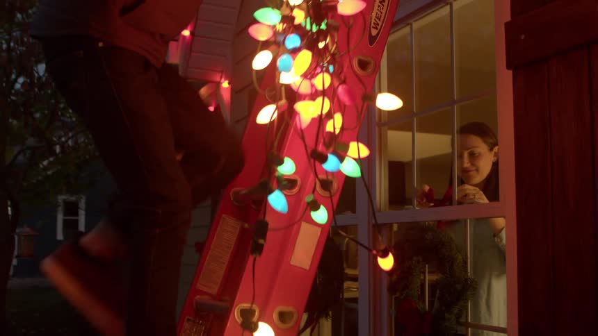 Woman inside the house installing a garland and outside a man descending a staircase with christmas lights