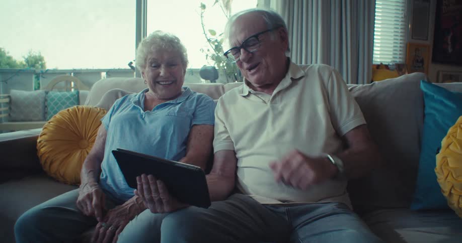 Elderly couple sitting on couch happily video calling on tablet