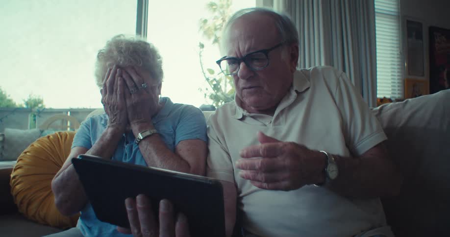 An elderly couple sits on a couch beside the window at midday with the man holding a tablet as they both watch in disappointment and sadness