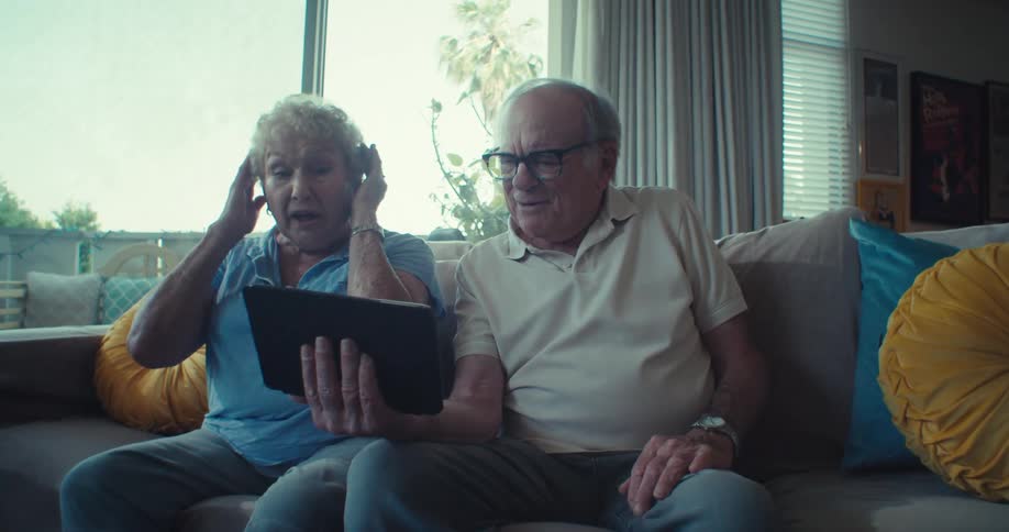 Elderly couple video calling and reacting warmly while sitting on a cozy living room couch