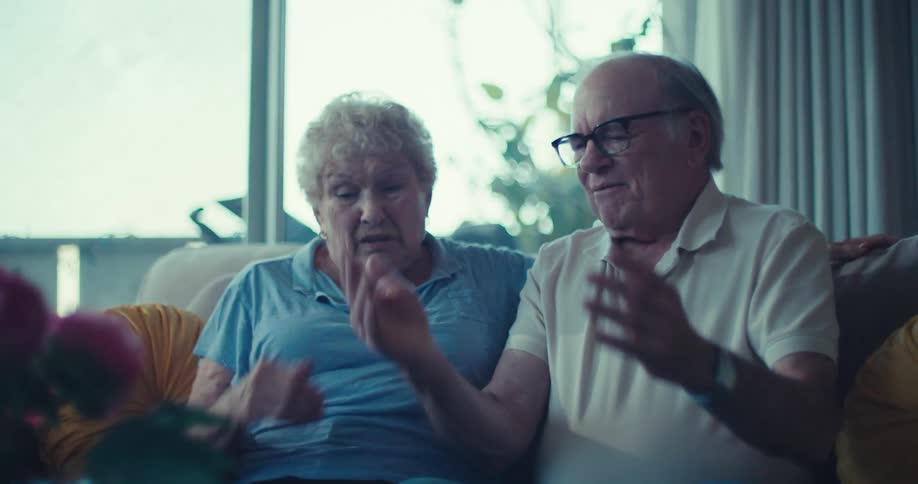 An elderly couple sitting together on a sofa looking at and interacting with a tablet device they appear to be engaged in a conversation possibly discussing what they are seeing on the screen reflecting a moment of shared experience and connection