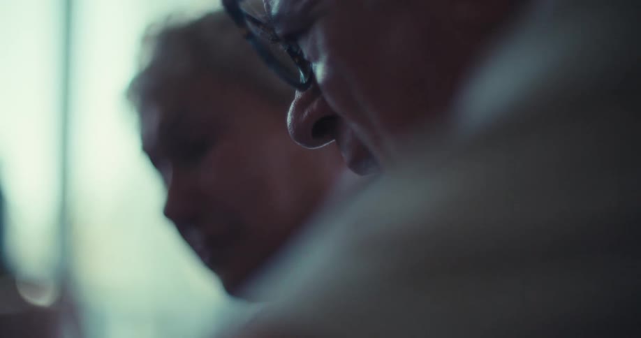 An elderly man wearing glasses talks and shows a tablet screen to a woman sitting beside him indoors