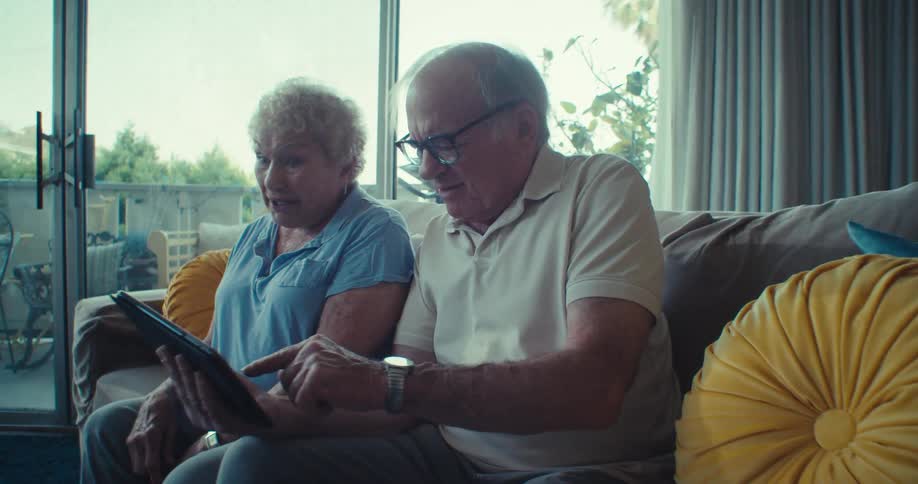 Elderly man and woman sitting on a sofa in the living room and interacting with a tablet