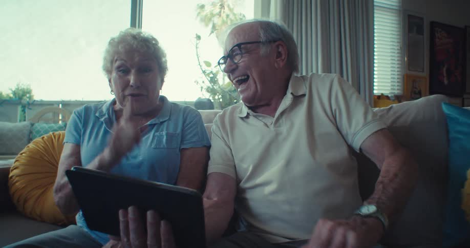 An elderly couple sits on a couch in a cozy living room sharing a laugh as one holds a tablet