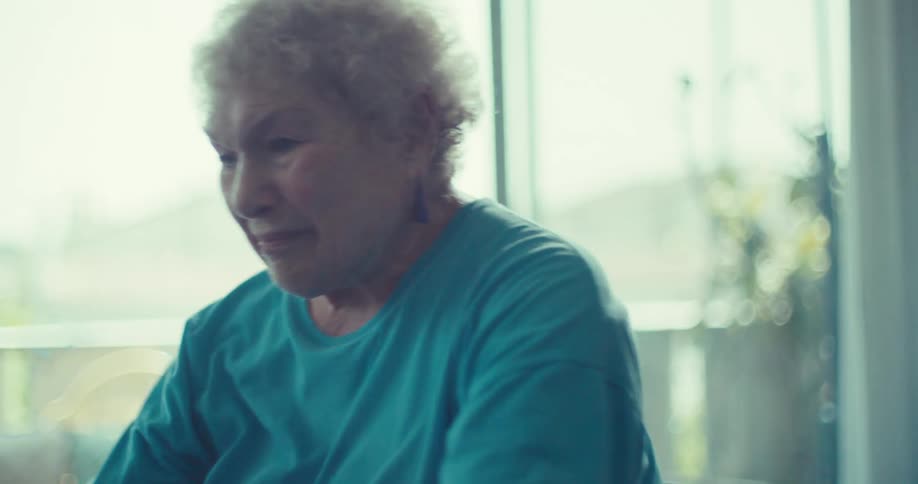 An elderly woman with short curly white hair wearing a blue tshirt she is initially seen giving someone a highfive smiling and appearing happy towards the end of the clip her expression becomes more somber or serious as she looks downwards
