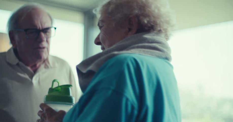 An elderly man with gray hair wearing glasses and a lightcolored polo shirt talking to an elderly woman with white curly hair who is dressed in a blue top and sitting on a stationary bike they appear to be in a bright room possibly a living room