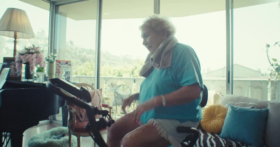 An elderly woman rides a stationary bike in a cozy sunlit living room