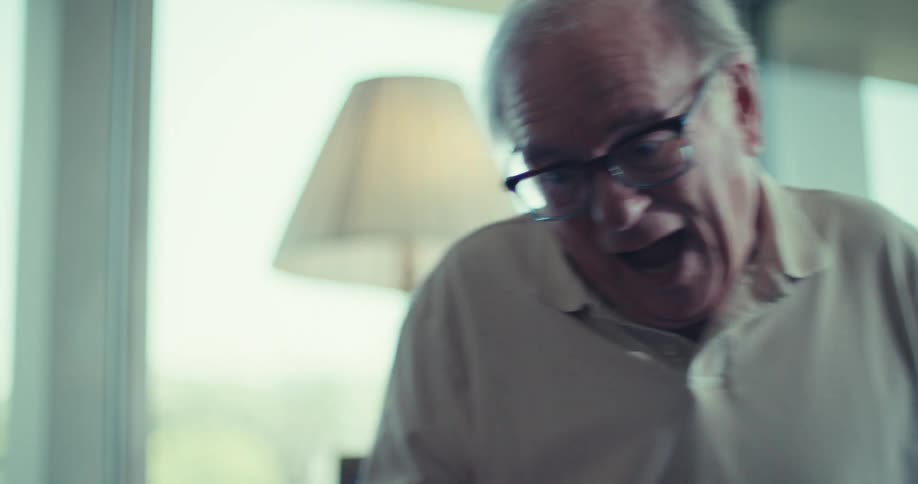 A smiling elderly man with glasses has an animated and joyful conversation with someone off screen in a brightly lit room