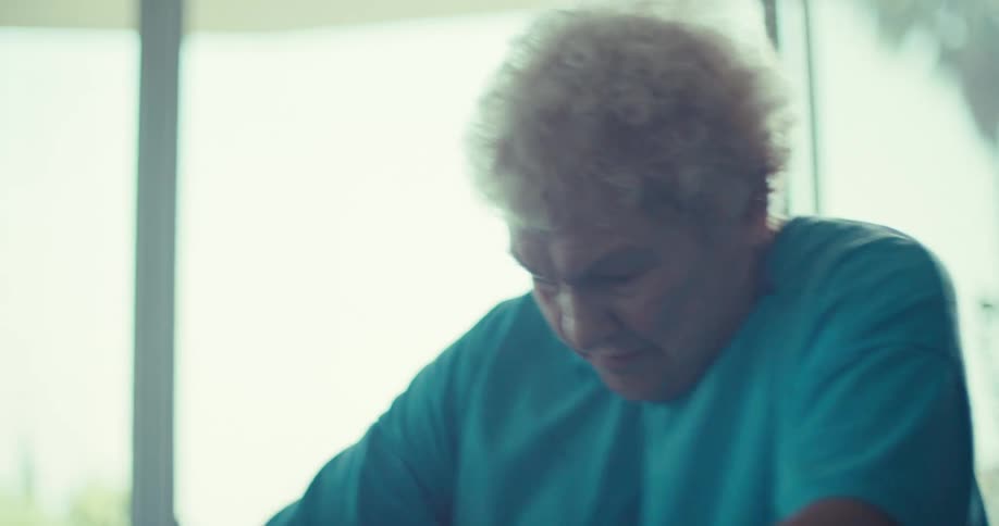An elderly woman with white curly hair wearing a teal tshirt exercising on what appears to be a stationary bike she is seen pedaling slowly and steadily her face showing concentration and effort