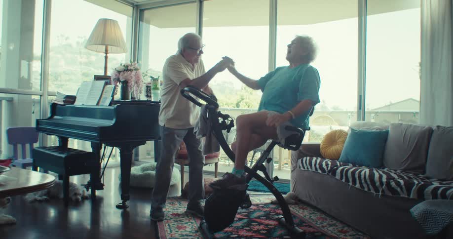 Two elderly individuals in a sunlit living room one person is on a stationary bike while the other stands beside them encouraging and highfiving them as they exercise