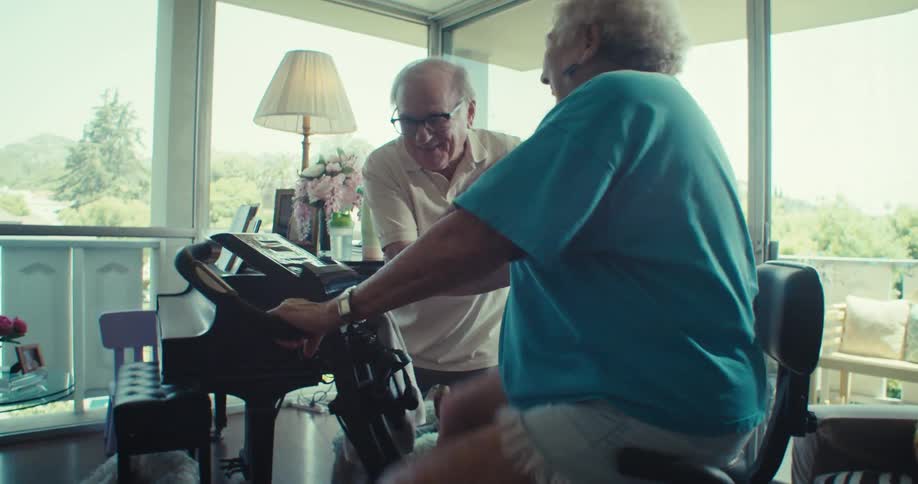 Elderly man encouraging elderly lady who is cycling on an exercise bike in living room