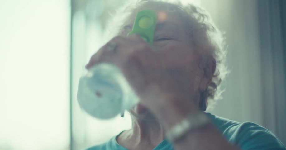 A closeup of an elderly woman with white curly hair wearing a blue top drinking from a green and white water bottle she takes several sips appearing refreshed and satisfied
