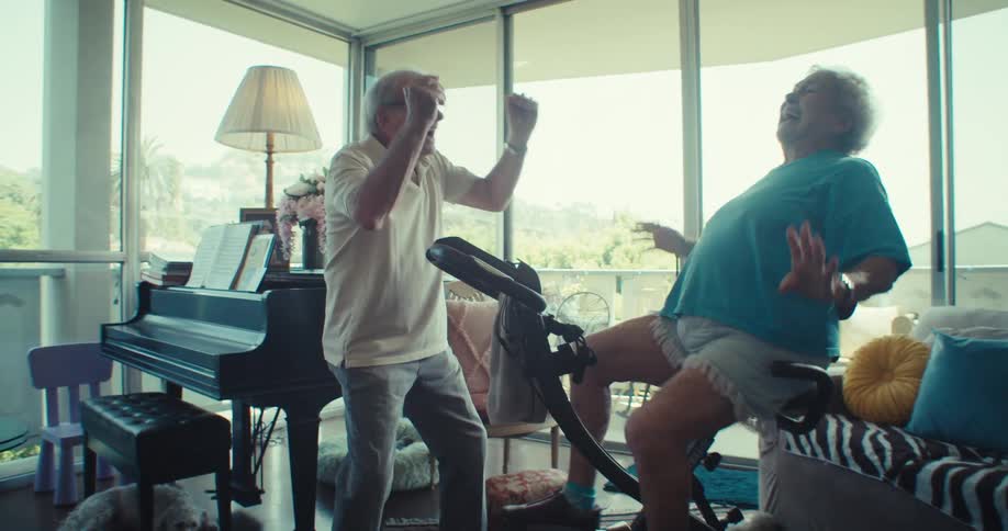 An elderly couple in what appears to be a brightly lit living room with large windows the woman is on an exercise bike laughing while the man enthusiastically encourages her eventually highfiving her