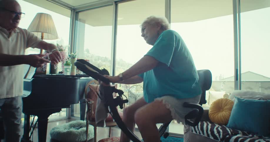 Elderly man supports his wife as she exercises on a stationary bike in their cozy living room