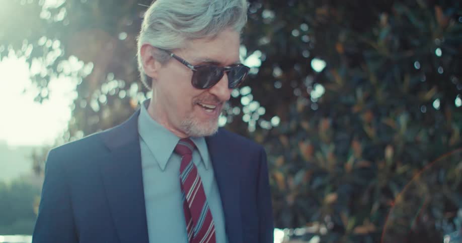 An older businessman with grey hair and a beard wearing sunglasses and a suit with a striped tie who smiles and takes off his sunglasses while walking he looks directly at the camera then down as if reading something