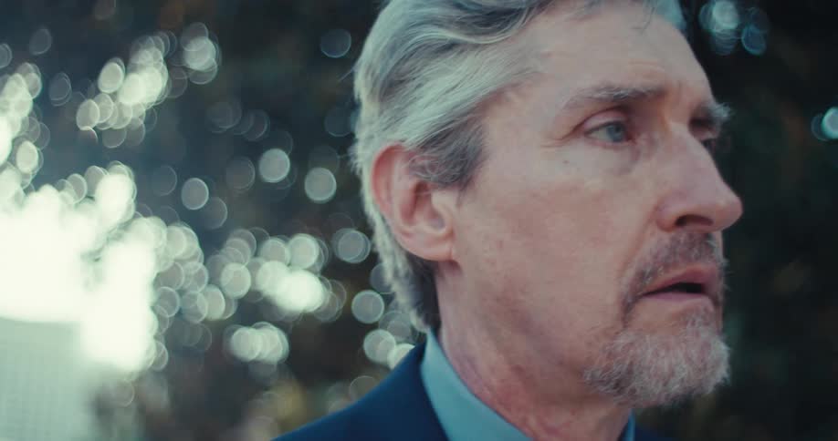 A stylish man with grey hair and a tie talks on a phone looking concerned or surprised while standing outdoors the background suggests an urban setting with blurred buildings and natural light filtering through trees