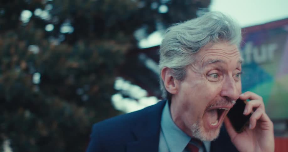 An older man with gray hair dressed in a suit and tie initially engrossed in his phone he then takes a call and his expression dramatically shifts to one of excitement and joy culminating in him throwing his hands up in triumph while walking away