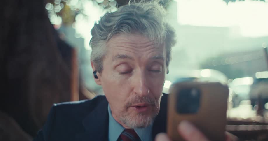 A middleaged man with graying hair and a beard wearing a suit and what appears to be an earpiece intently looking at his phone he seems surprised or engrossed by something on the screen as his mouth opens slightly