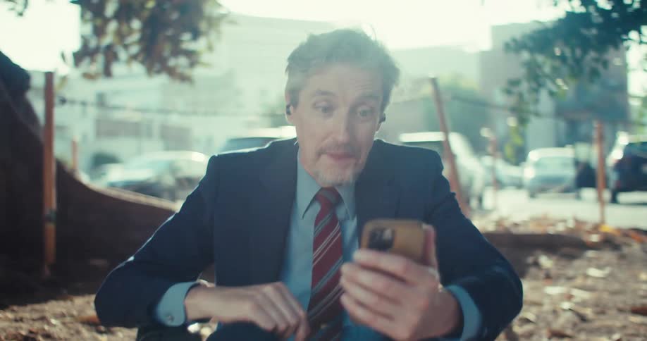 A man in a suit sits on a curb near a parking lot smiling and raising a hand in celebration while taking a call beside a closed briefcase