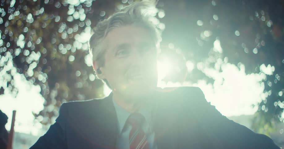 A businessman in a suit and tie wearing earbuds smiles and looks upwards with an expression of enjoyment or contemplation the bright sunlight filters through the trees in the background creating a dappled lens flare effect around him