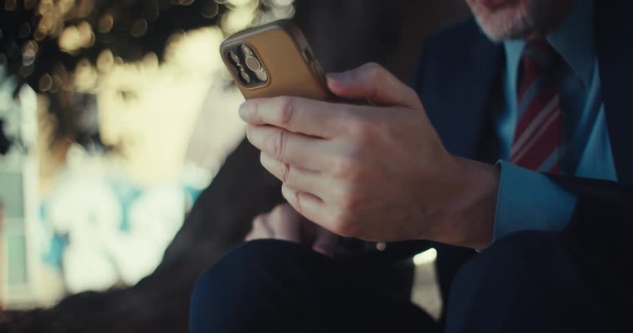 A man in a suit reacts with excitement while looking at his phone outdoors