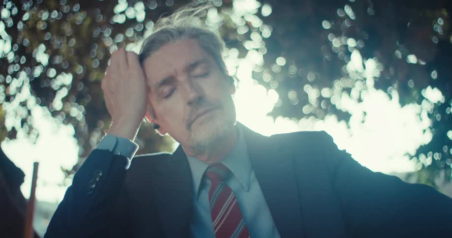 Older businessman sitting under a tree on a windy day listens to music using his smartphone and earbuds