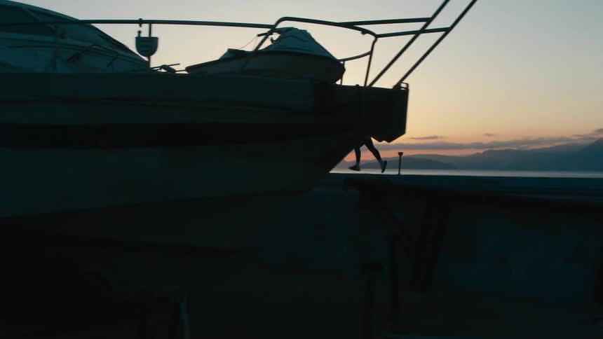 A docked boat sits in silhouette against the soft glow of a sunset as a female runs on deck