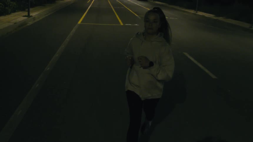 A woman with dark hair tied back wearing a lightcolored hoodie and dark leggings jogging on a paved surface marked with yellow lines it appears to be nighttime and the area is dimly lit suggesting an outdoor track or a quiet road