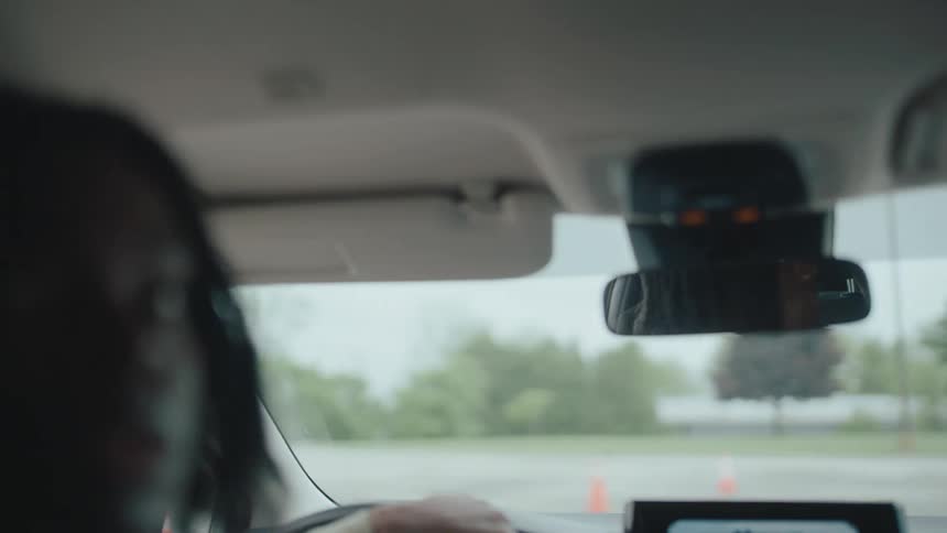 A young woman steers a car through a parking lot while navigating around orange traffic cones visible beyond the windshield