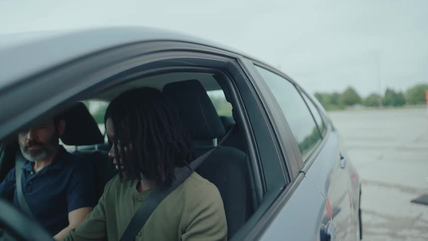 A person with long dark hair possibly dreadlocks in the drivers seat of a car looking forward with a serious expression another person is visible in the passenger seat though their face is partially obscured