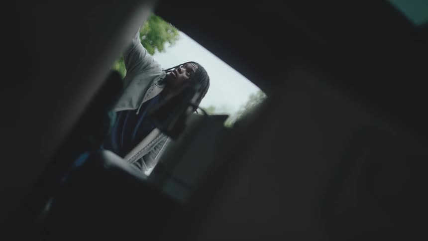 A person with long dark hair wearing a lightcolored top reaching into the back of a car from an external perspective they appear to be retrieving or placing an object into the vehicles trunk or back seat before closing it