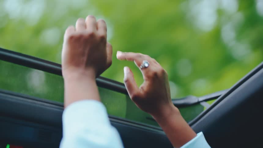 A persons hands reach up to close the sunshade on a cars sunroof with blurred green foliage visible outside